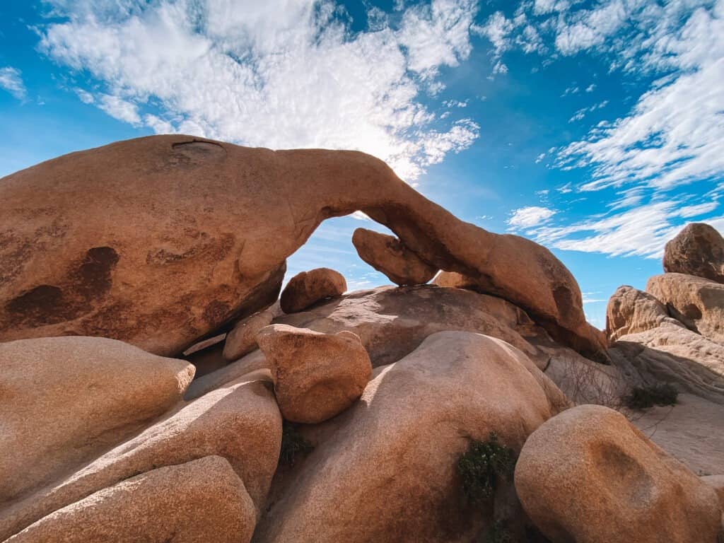 A long curved arch rock against a bright blue sky and surrounded by boulders in Joshua Tree National Park.