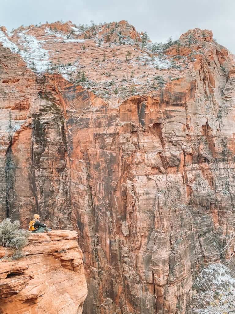 a hiker sits at the edge of a vast red rock canyon at Zion Canyon Overlook in Zion National Park.